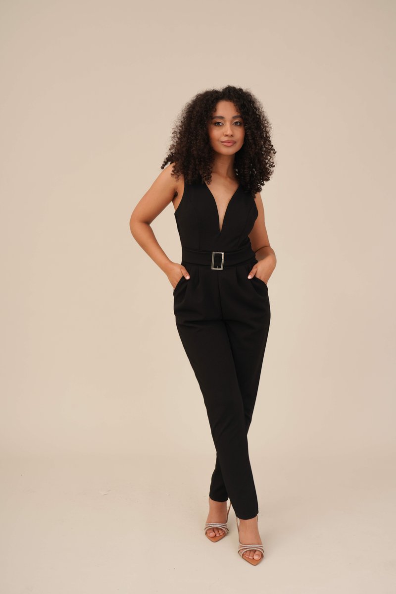 Μαύρο φόρεμα με βαθύ V ντεκολτέ, ζωνάκι στη μέση και πλευρικές τσέπες. Το μοντέλο φορά διαφανείς sandals με τακούνι. Λείο ύφασμα και κομψή εφαρμογή.