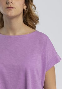 Top corto a maniche corte di colore viola chiaro, con scollatura ampia e un tessuto morbido e testurizzato. I dettagli delle cuciture sono minimi, esaltando il design informale.