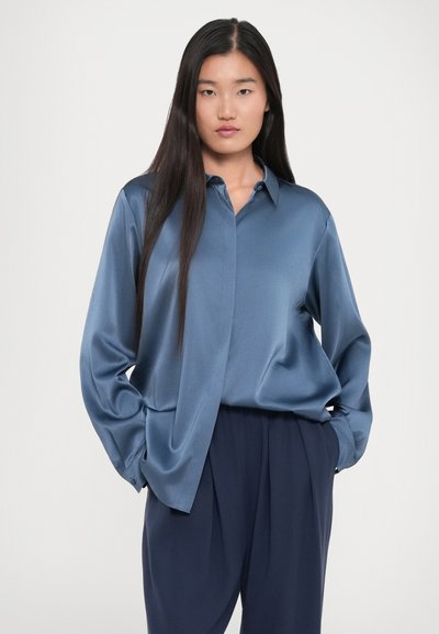 Joven mujer con cabello largo y oscuro viste una camisa de satén azul metida dentro de unos pantalones de pinzas navy, de pie con las manos en los bolsillos contra un fondo simple.