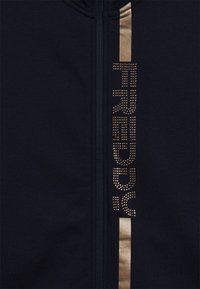 Tessuto scuro con una striscia verticale metallica dorata accanto a una zip, con la parola "TRUE" scritta in piccole borchie dorate.