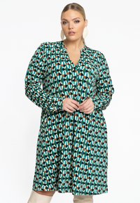 Robe verte avec un motif géométrique en teal, orange, crème et noir. Dispose d'un décolleté en V, d'une fermeture par bouton et de manches longues.
