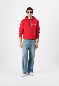 Rød bomull hettegenser med frontlomme og snorer; har "TOMMY HILFIGER"-logo. Brukt sammen med lyseblå jeans og hvite sneakers.