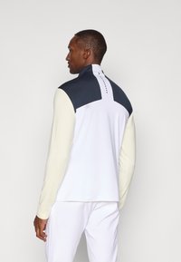 Camisola branca de mangas longas com realces azul escuro nos ombros e gola alta. Apresenta uma textura suave e um design com caimento traseiro. Detalhe de logo discreto.