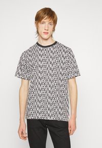 Camiseta blanca con el logo negro de "Levi's" repetido en patrones rectangulares, cuello redondo y mangas cortas. Se lleva con pantalones negros.
