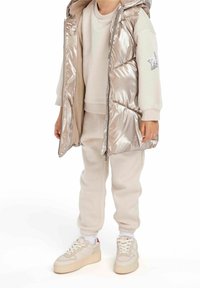 Chaleco plateado acolchado sobre una sudadera beige y pantalones jogger a juego, combinados con zapatillas claras con detalles rojos y suelas texturizadas.