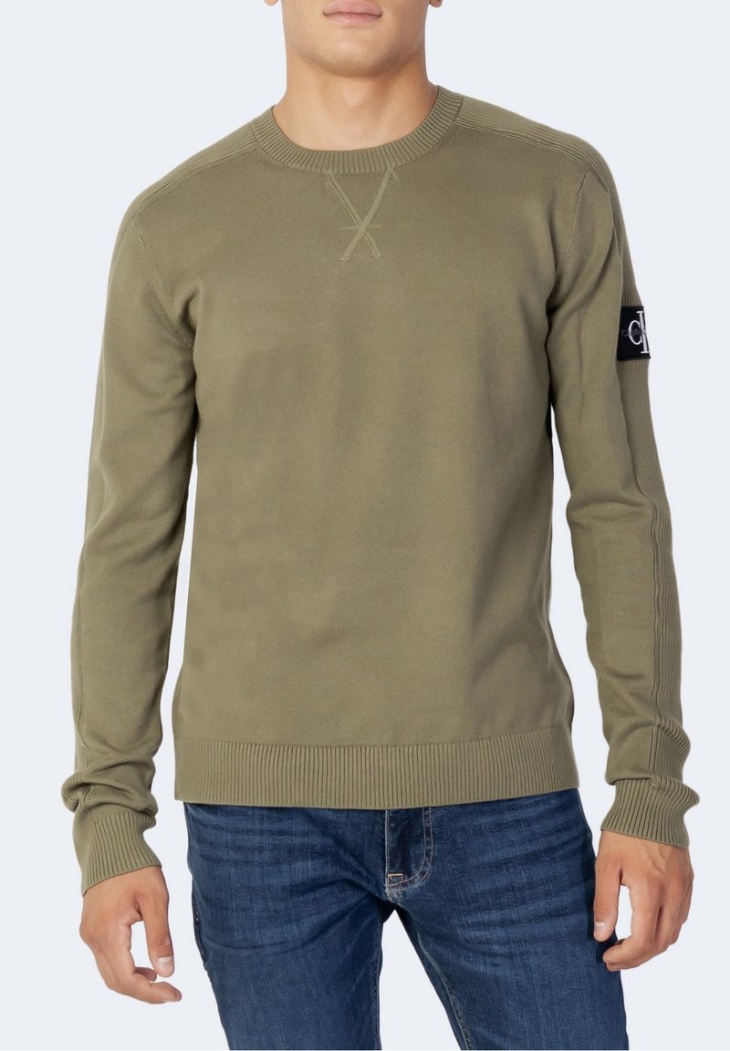 Homme portant un pull vert olive à manches longues avec col rond, poignets et ourlet côtelés, associé à un jean bleu, debout devant un fond uni.