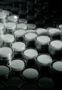 Des contenants en plastique transparent remplis de crème blanche, disposés de manière dense dans un cadre sombre, avec des couvercles ronds et plats surmontés d'un petit point.