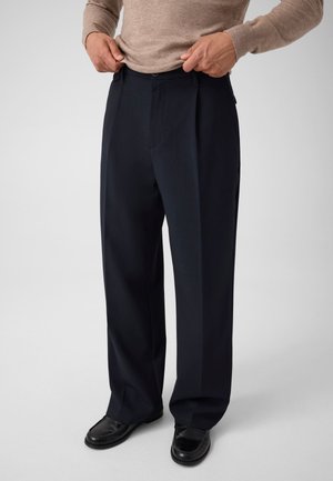 Personne ajustant la taille d'un pantalon noir ample porté avec des chaussures noires et un haut beige à manches longues sur un fond uni.