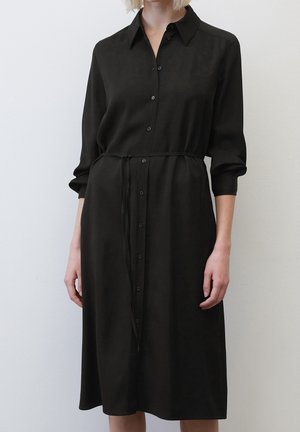 Kobieta ubrana w czarną sukienkę midi na guziki z długim rękawem, kołnierzykiem i cienkim paskiem przewiązanym w talii, stoi na tle jednolitego tła.