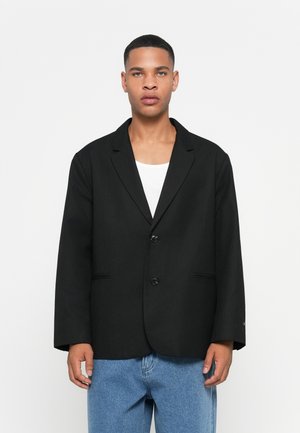 Blazer nero con una vestibilità rilassata, due bottoni, rever a taglio e due tasche laterali. Indossato sopra una camicia bianca e abbinato a jeans blu.