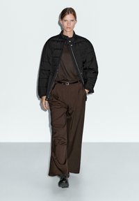 Woman walking in black puffer jacket, dark brown top and wide-leg trousers, with black belt and shoes against plain light background.