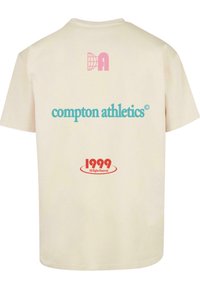 Kremkrāsas kokvilnas T-krekls ar rozā globusa logo un "Compton Athletics" teal krāsā, kā arī sarkanu "1999 Visas tiesības rezervētas" grafiku.