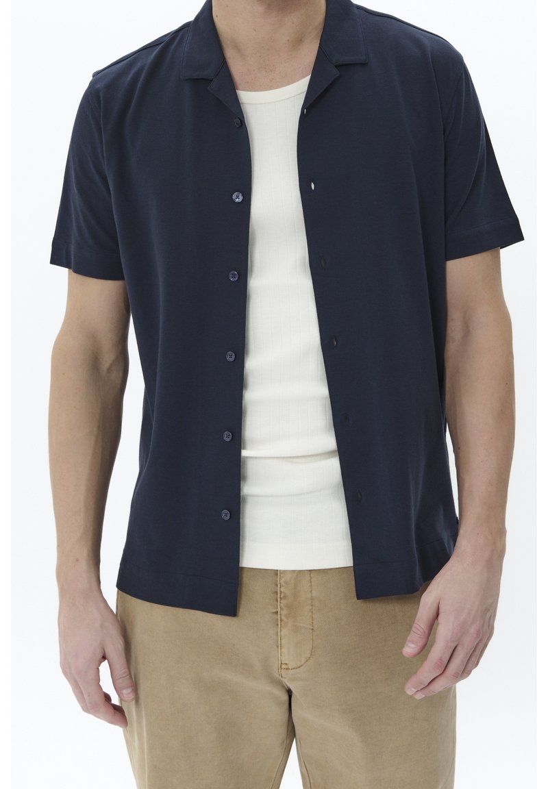 Marinblå kortärmad skjorta med campkrage, framknäppning och slät textur, bärs över en vit ribbad linne och matchas med beige byxor.
