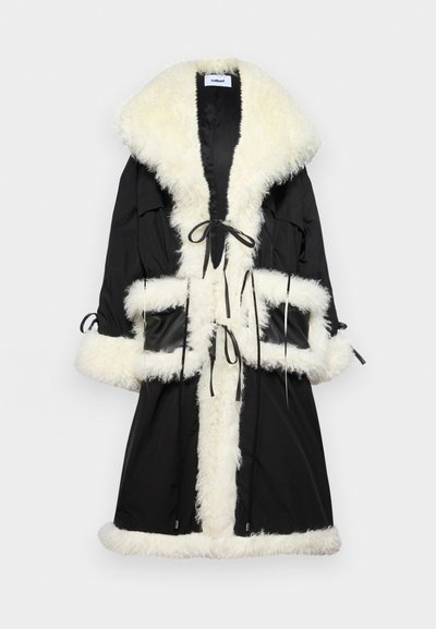 Zwarte buitenkleding met een nepbontkraag en -afwerking in crème. Voorzien van trekkoordsluitingen en twee voorzakken. Het materiaal lijkt soepel en warm.