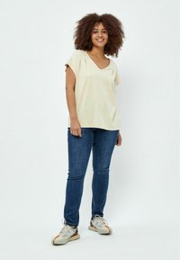 Cremfarbendes V-Ausschnitt-Shirt mit kurzen Ärmeln, kombiniert mit dunkelblauen Jeans und mehrfarbigen Sneakers mit Schnürsenkeln und strukturierten Sohlen.