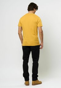 Tricou cu guler rotund de culoare muștar, siluetă ajustată, asortat cu pantaloni negri și bocanci bej, prezentat din spate pe un fundal deschis.