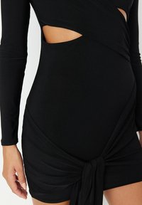 Abito aderente nero con maniche lunghe, caratterizzato da dettagli con tagli e un design con nodo in vita. Realizzato in tessuto liscio con una leggera lucentezza.
