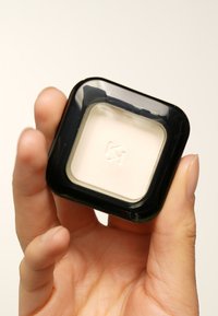 Conteneur noir carré avec un couvercle transparent, contenant un produit de crème légère à l'intérieur, embossé avec un logo. Main tenant le conteneur.