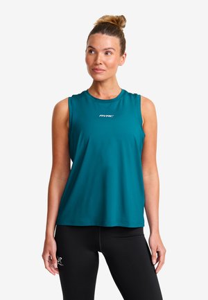 Woman in a teal sleeveless top and black leggings standing against a plain white background, looking slightly to the side.
