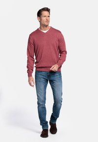 Rode V-hals trui met lange mouwen, gedragen over een witte blouse, gecombineerd met blauwe spijkerbroeken en bruine suède laarzen. Eenvoudig, casual ontwerp.