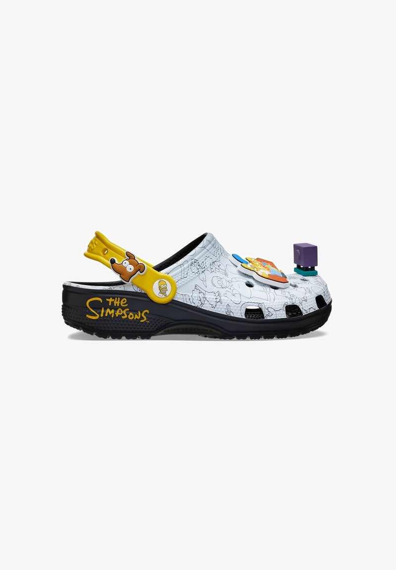 Weiße Crocs mit Cartoon-Designs, einem gelben Riemen, schwarzem Boden und verschiedenen bunten Charm-Zubehörteilen. Detail mit "Die Simpsons"-Marke.