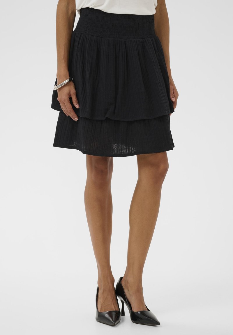 Femme portant une jupe noire à plusieurs couches longueur genou, un haut blanc, un bracelet argenté et des chaussures à talons hauts noires à bout pointu, debout devant un fond uni.