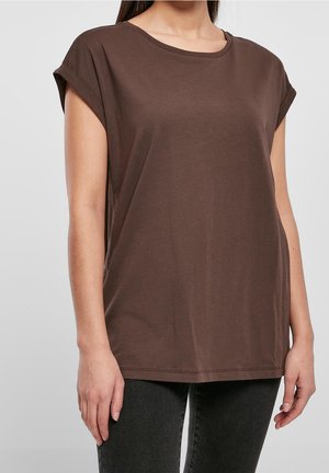 Femme portant un t-shirt marron ample à manches courtes et un pantalon foncé, debout devant un fond gris clair uni.