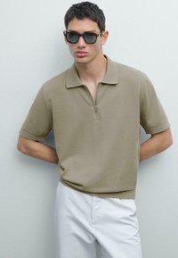 Polo tričko s krátkým rukávem v světlém taupe s zipovým límečkem; v kombinaci s bílými kalhotami. Má hladkou texturu a jednoduchý design.