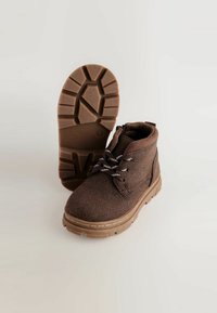 Brūni, vidukļa zābaki, kas izgatavoti no teksturēta auduma ar mīkstu zoli, ar apaļām auklām un zīmējumu gumijas zoli labākai saķerei.