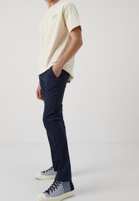 Béžové bavlněné tričko s krátkými rukávy a modrým logem, ve spojení s tmavě modrými kalhotami a vícebarevnými teniskami s bílou podrážkou.