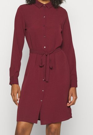 Woman wearing a long-sleeve, button-front burgundy dress with a tied waist belt, shown from shoulders to knees against a plain background.