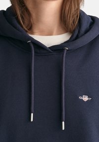 Primo piano di una persona che indossa un hoodie blu navy con lacci e un piccolo logo ricamato sul petto sopra una camicia bianca.