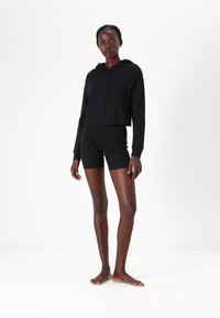 Čierna skrátená mikina na zips s kapucňou a prednými vreckami, skombinovaná s čiernymi priliehavými šortkami. Model bez topánok na jednoduchom pozadí.