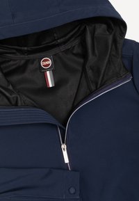 Temno modra jakna s policom odprtim srebrnim zadrgo, kapuco in črto notranjo podlogo, ki prikazuje etiketo Colmar Originals in trirazredno tkanino oznako.