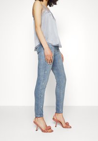 Lichtgrijze satijnen tanktop met kantafwerking, gecombineerd met blauwe skinny jeans met gele geborduurde details en roze haksandalen.