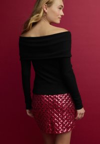 Top nero a costine off-the-shoulder abbinato a una mini gonna rossa scintillante con paillettes, caratterizzata da un motivo a diamante. Posizionata su uno sfondo rosa.