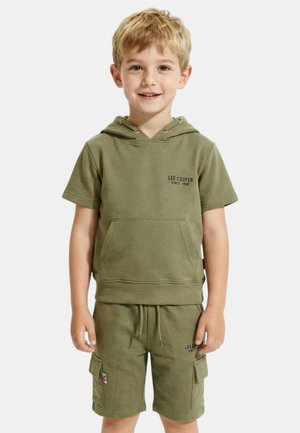 Garçon blond souriant, portant un sweat à capuche à manches courtes et un short cargo assortis de couleur vert olive avec le logo "Lee Cooper" sur les deux.