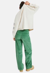 Vaaleanbeige halkaistu huppari, jossa on kukkakuvio hihassa, yhdistettynä vihreisiin samettihousuihin ja ruskeisiin, paksupohjaisiin saappaisiin.