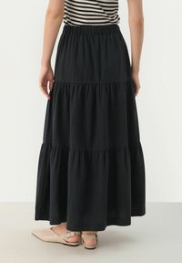Svart, lager-på-lager maxi kjol gjord av lättviktig tyg med elastisk midjeband. Stylad med krämfärgade slingback-skor. Bakvy.