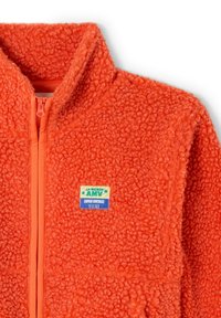 Jasnopomarańczowa kurtka polarowa z przednim zamkiem błyskawicznym i małą prostokątną naszywką z napisem "La Maison AMV Super Vintage" na piersi.