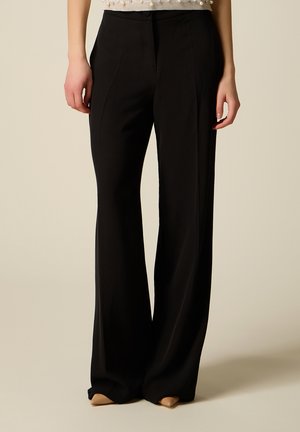 Kobieta w czarnych szerokich spodniach z wysokim stanem, zestawionych z beżowymi butami z noskiem w szpic na neutralnym tle.