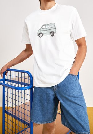 Mand iført hvid t-shirt med gråt varevognsprint og blå denimshorts, der læner sig op ad en blå metalvogn med træhylder.