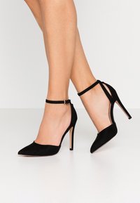 Černé semišové vysoké podpatky s špičatou špičkou a kotníkovým páskem, s jemným koženým povrchem a stiletto podpatkem.
