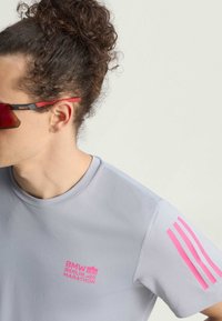 Osoba s kovrčavom kosom u punđi nosi crvene sportske sunčane naočale i sivi majicu iz Berlinskog maratona s ružičastim logom i prugama na rukavu.