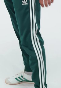 Grüne Sporthose mit drei weißen Streifen an den Seiten, kombiniert mit weißen Sneakers mit grünen Akzenten und einer braunen Sohle.