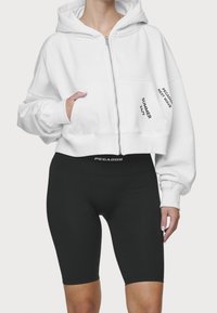 Sudadera blanca recortada con cremallera y bolsillos frontales, con texto negro. Combinada con pantalones cortos de ciclismo negros de cintura alta. Tejido suave.