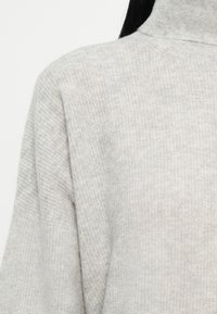 Svetrovka iz svetlo sive pletenine s turtleneck ovratnikom, nošena, prikazuje rame in zgornji del prsnega koša, izdelana iz mehkega teksturiranega blaga.