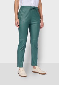 Pantalones de cuero sintético verde con una cintura elástica, bolsillos laterales y un diseño ajustado. Combinados con zapatillas de crema claro.