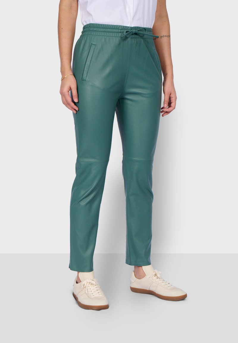 Pantalones de cuero sintético verde con una cintura elástica, bolsillos laterales y un diseño ajustado. Combinados con zapatillas de crema claro.