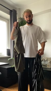 Camisa de manga curta verde-oliva numa mão e camisa preta riscada na outra, combinadas com calças pretas, num espaço interior iluminado.